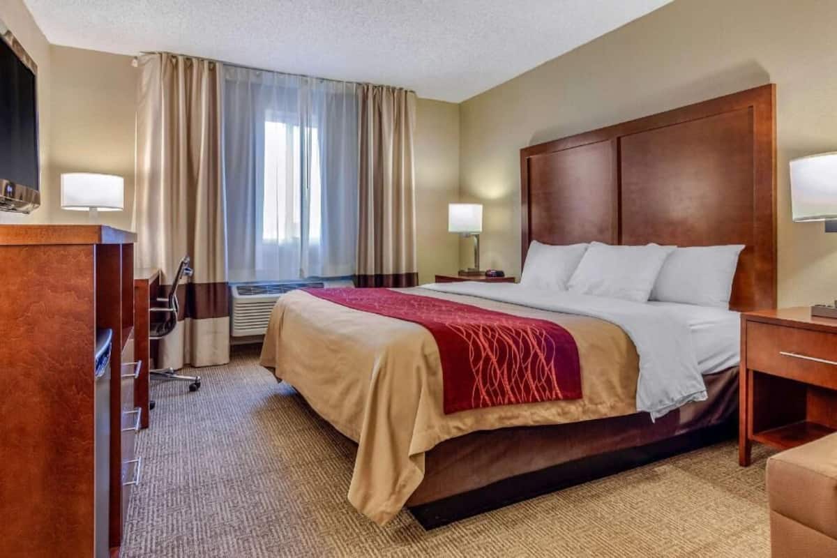 A hotel room with a bed, bedside tables, a desk, a TV, and a window with curtains.