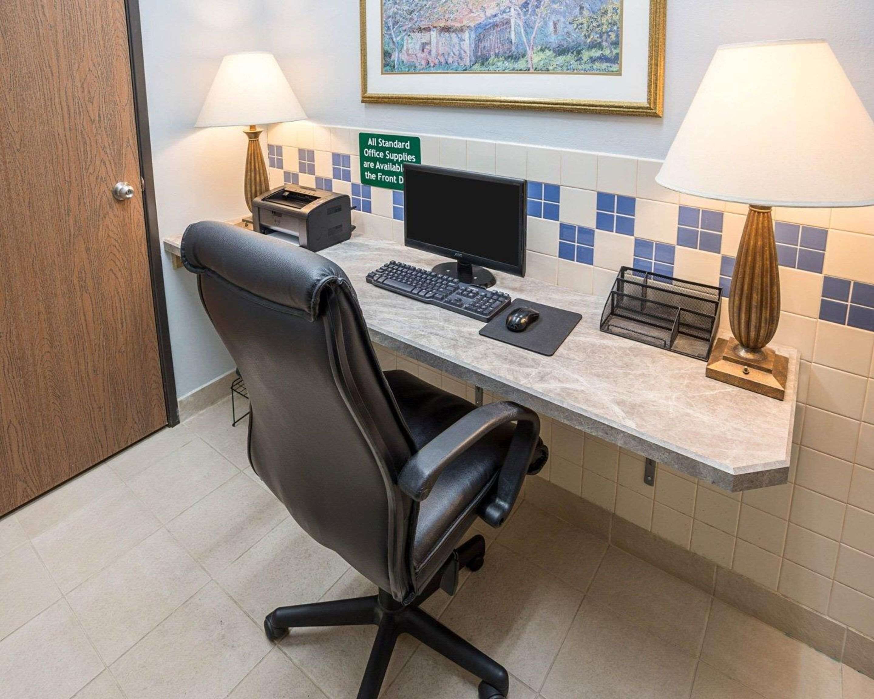 Oficina de hotel con escritorio, computadora, silla, lámparas y un cartel que indica la disponibilidad de material de oficina estándar.