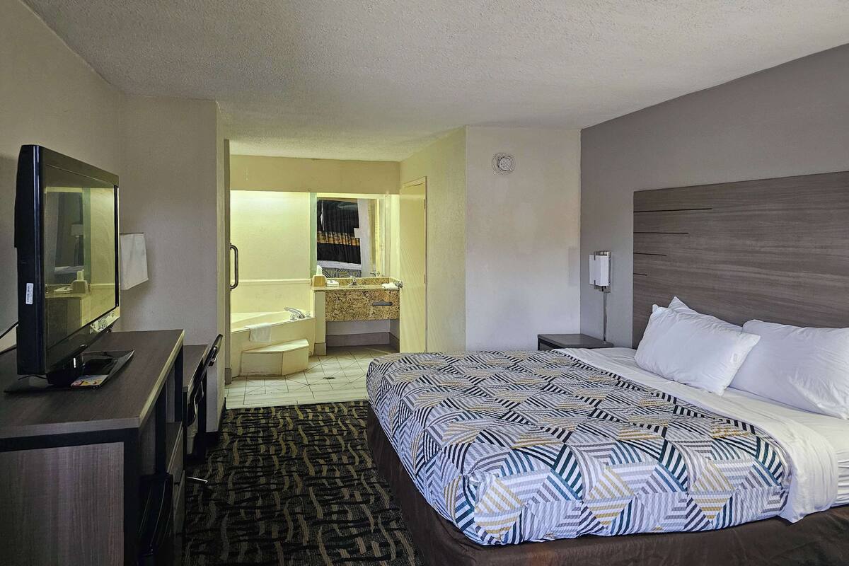 A hotel room with a bed, a desk, a television, a bathroom with a sink and mirror, and a carpeted floor.