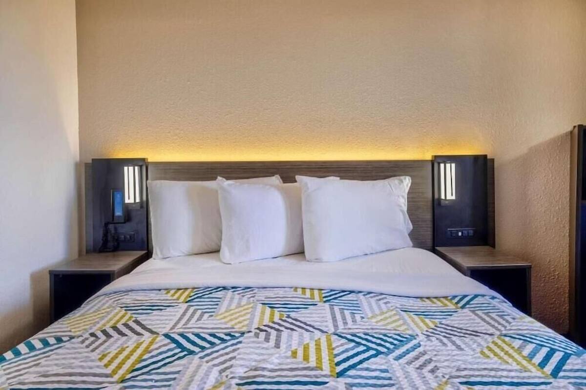 A hotel room with a bed, white pillows, a patterned bedspread, and bedside lamps.