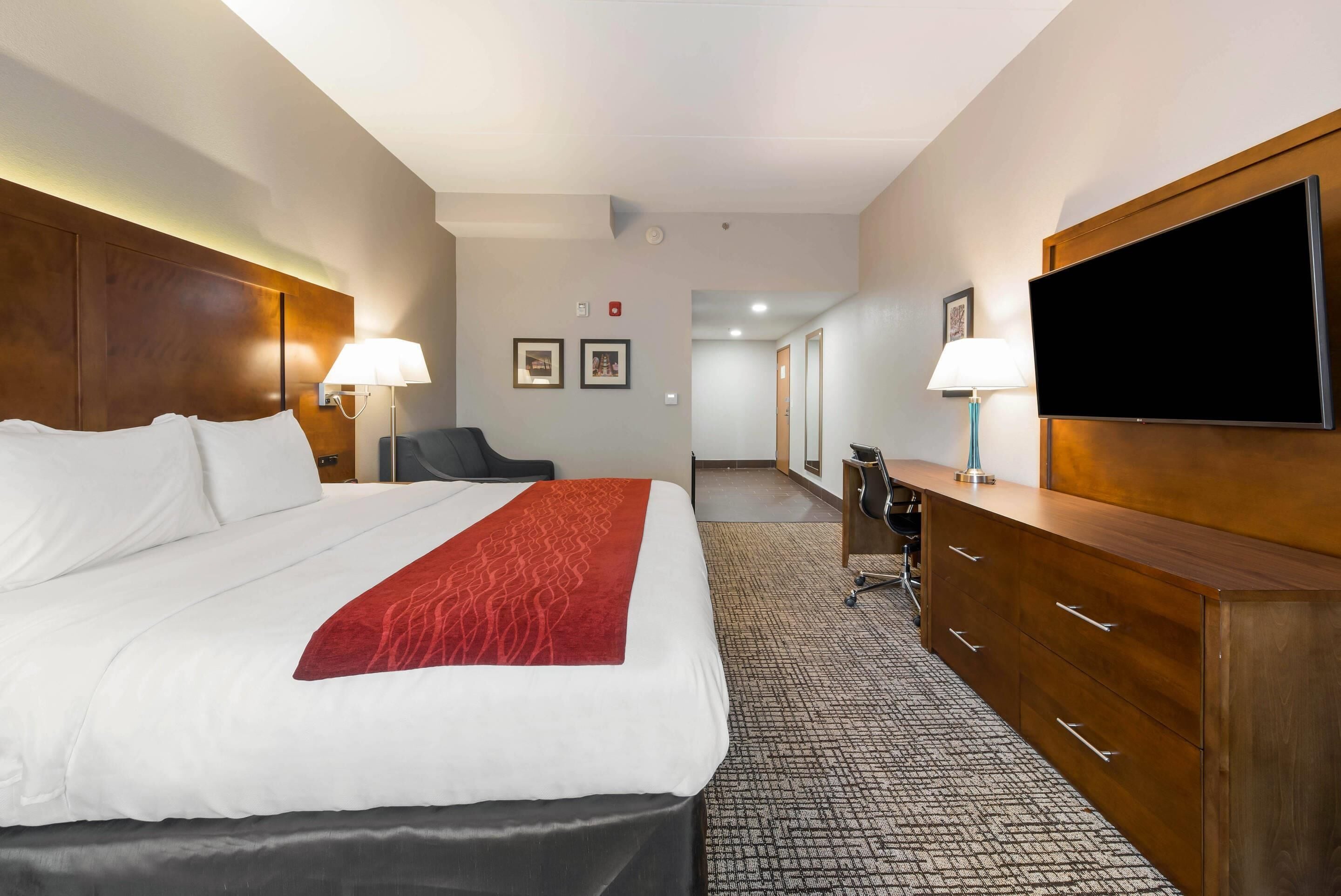 A hotel room with a large bed, a wooden dresser, a desk, a chair, and a television.