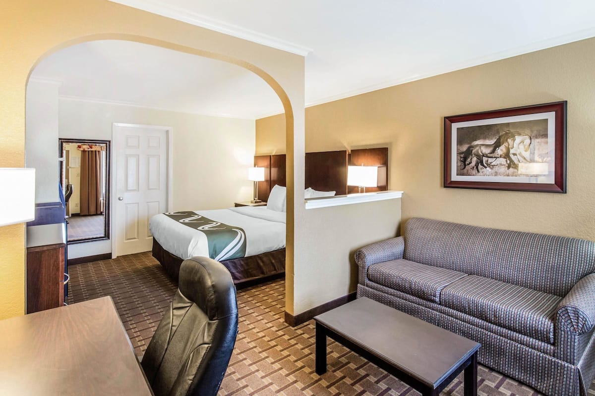 A hotel room with a bed, a sofa, a table, and a chair. There is a door leading to another room.