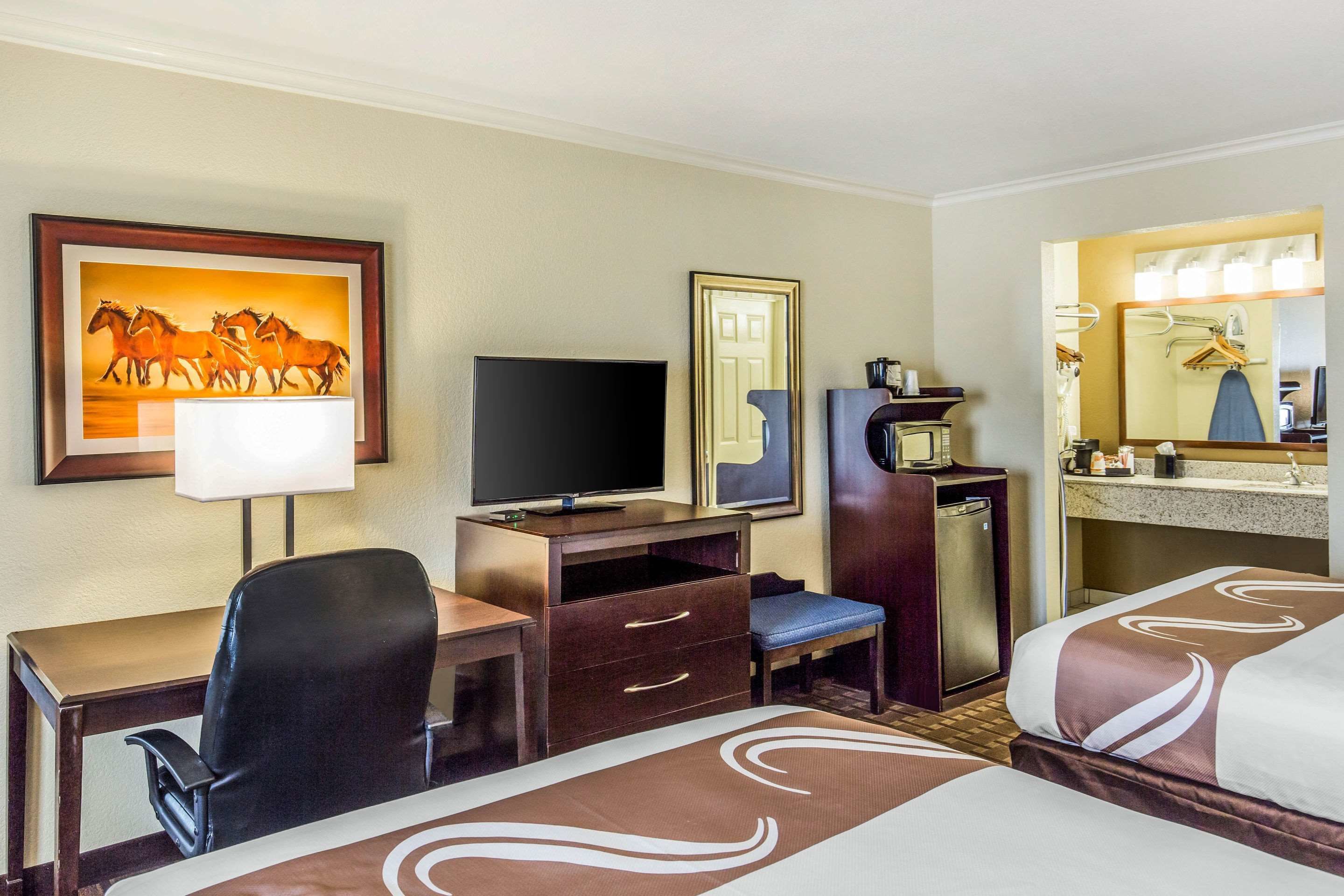 A hotel room with a bed, a desk with a chair, a television, a minibar, a mirror, and a painting of horses.