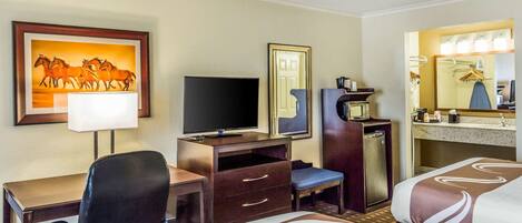 A hotel room with a bed, a desk with a chair, a television, a minibar, a mirror, and a painting of horses.