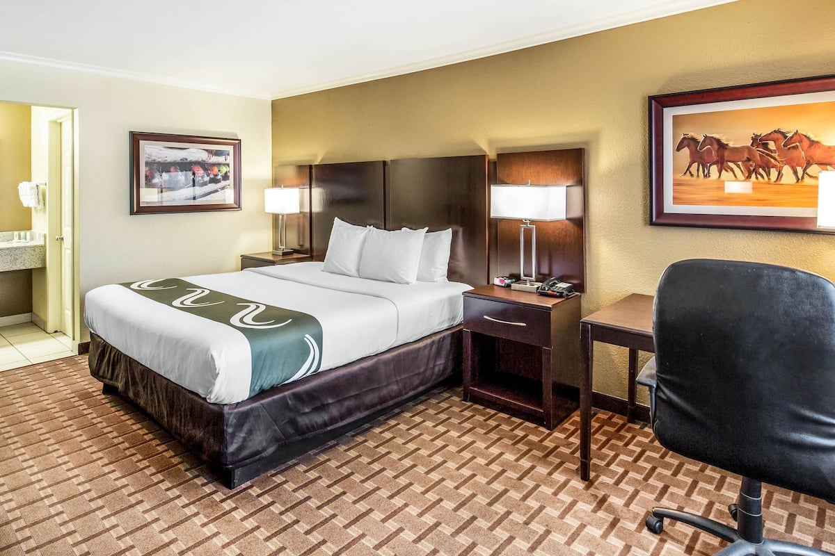 A hotel room with a large bed, a desk, two lamps, a chair, and artwork on the walls.