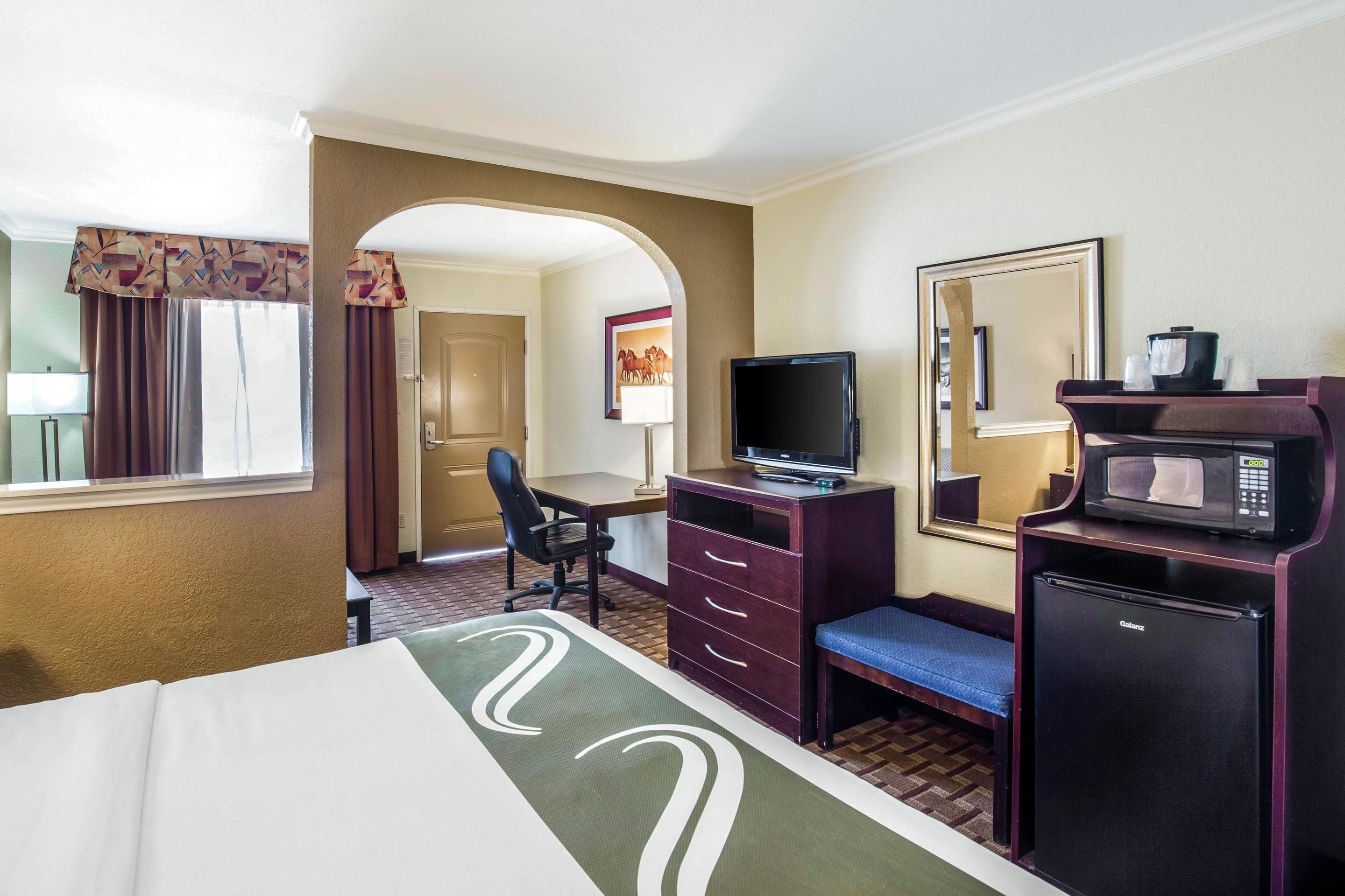 A hotel room with a bed, a desk with a computer, a television, a microwave, and a refrigerator.