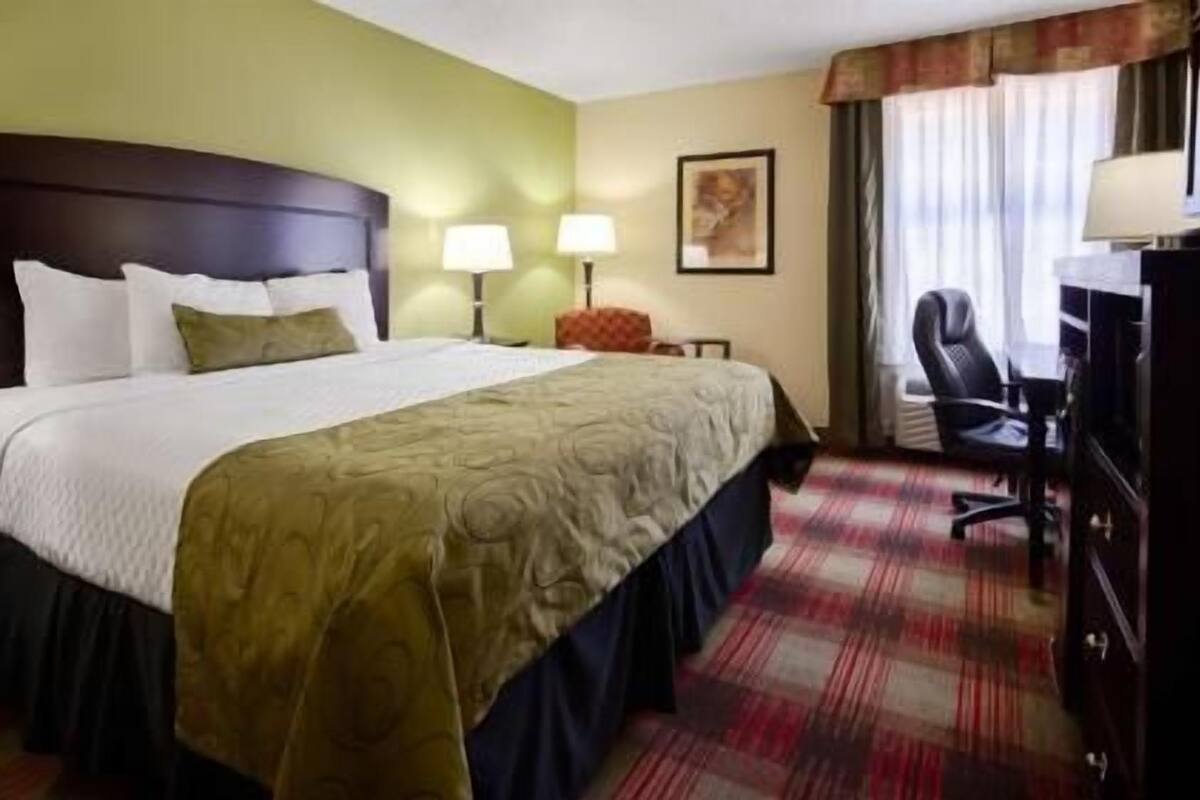 A hotel room with a bed, two bedside lamps, a desk with a chair, and a window with curtains.