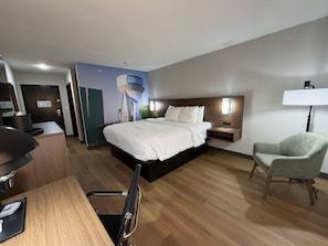 A hotel room with a bed, bedside table, lamp, chair, desk, and a wall with a poster. - Clarion Pointe Addison - Dallas North (Dallas)