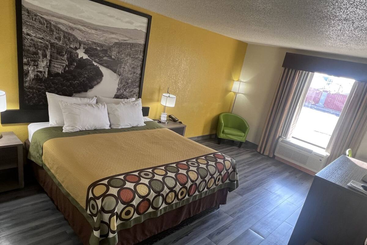 Een hotelkamer met een bed, nachtkastje, lamp, stoel, groot schilderij en een raam.