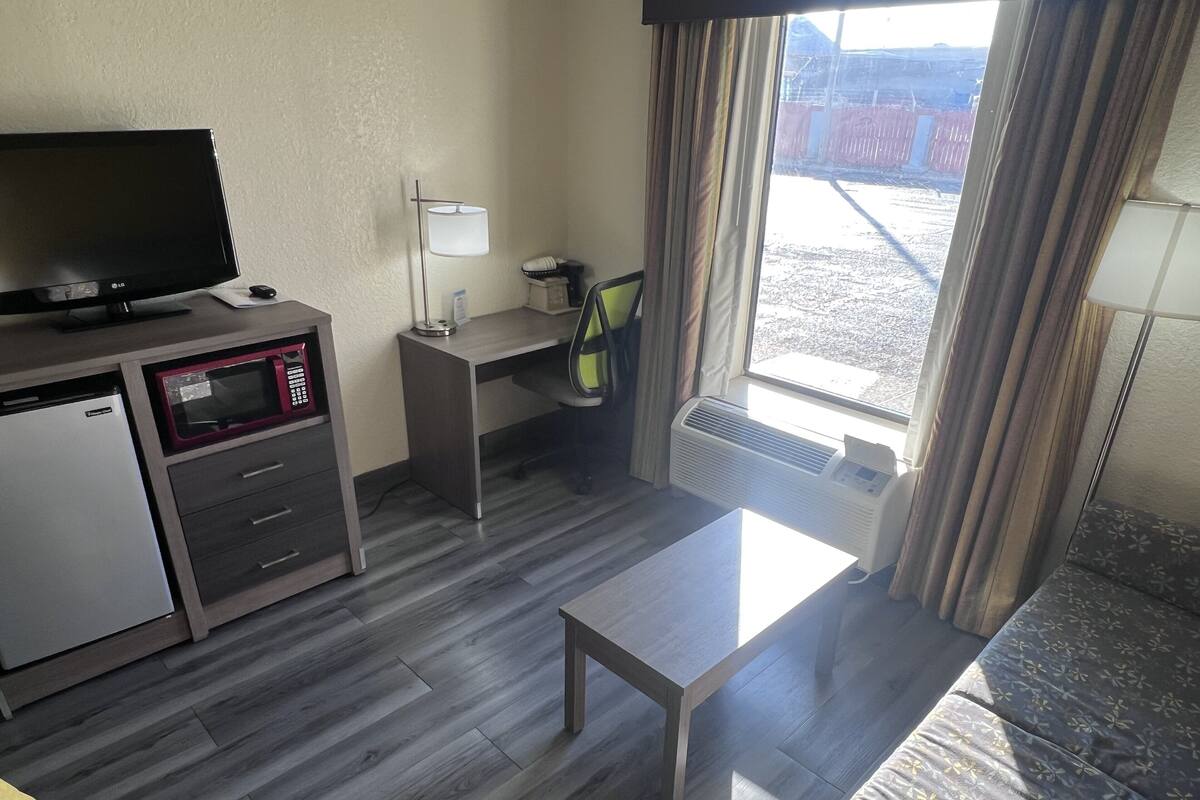 Een compacte hotelkamer met kitchenette, flatscreen televisie, bureau, stoel, bank en een groot raam met gordijnen.