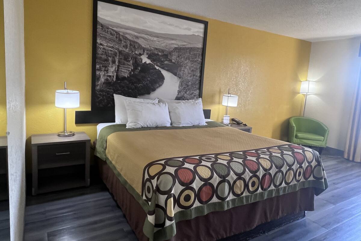 Een hotelkamer met een bed, twee bedlampjes, een groene fauteuil en een groot landschap op de muur.