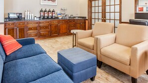 Un salon comprenant un canapé, un pouf et des fauteuils, un coin cuisine avec un four à micro-ondes et une machine à café, ainsi qu’une grande porte en bois.