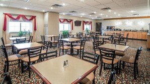 A hotel dining area with multiple tables and chairs, a kitchen in the background, and patterned carpet.