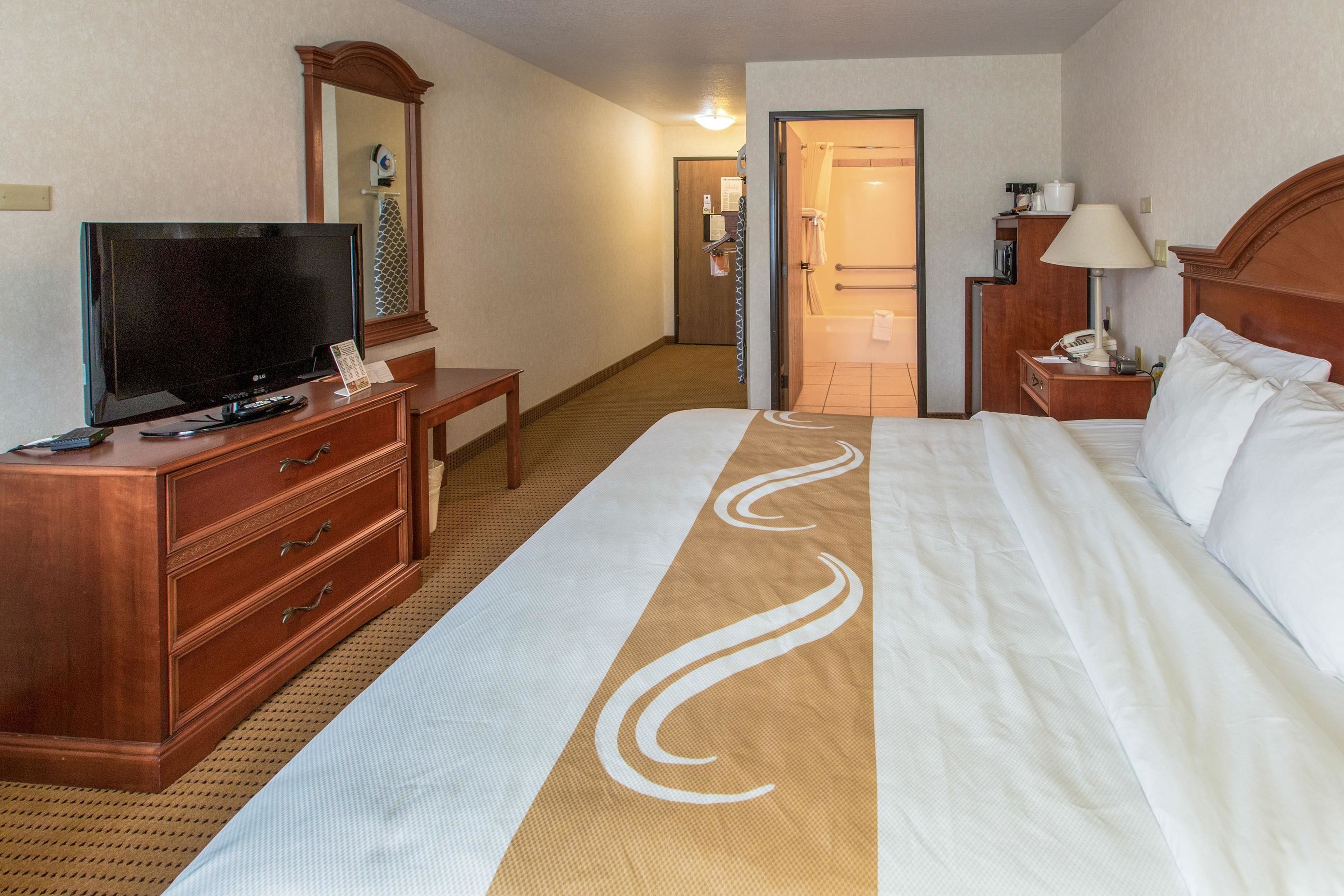 Una habitación de hotel con una cama grande, un televisor sobre una cómoda de madera, un espejo y un baño visible a través de una puerta abierta.