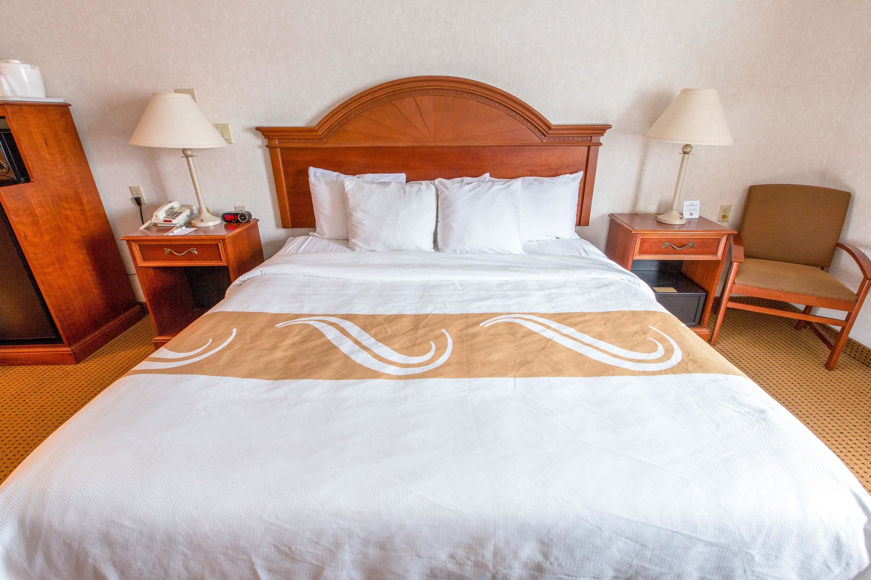 A hotel room with a large bed, two bedside tables with lamps, a wooden nightstand, a chair, and a wardrobe.