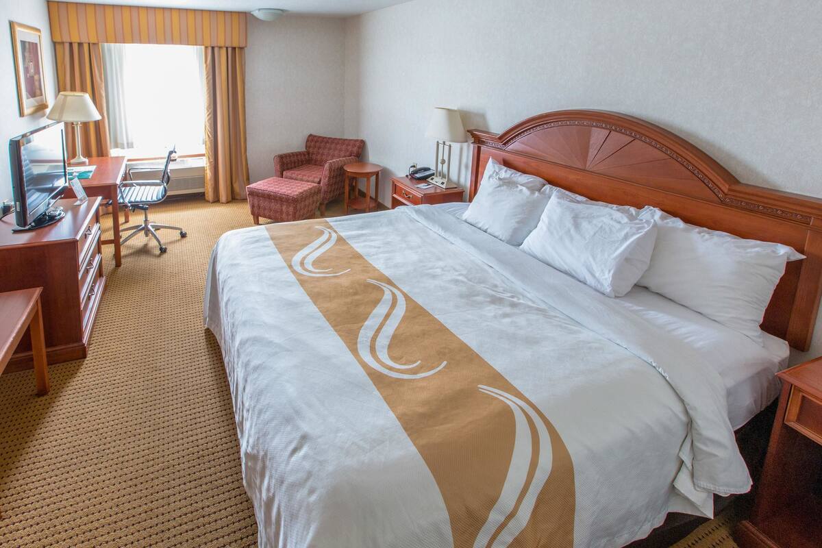A hotel room with a large bed, a desk with a computer, a chair, and a small table with a lamp.