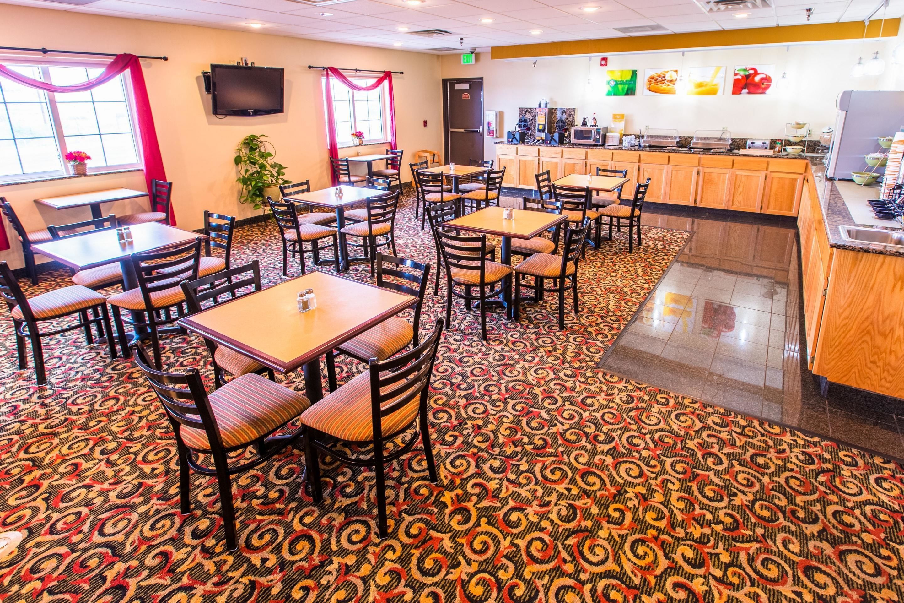 Un restaurante con alfombra estampada, mesas y sillas de madera, una barra con estación de bebidas y un televisor montado en la pared.