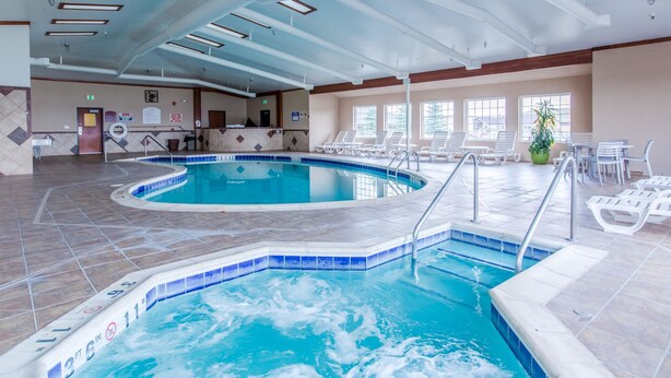 An indoor swimming pool with clear water, tiled flooring, and a sunlit area with chairs and a plant.