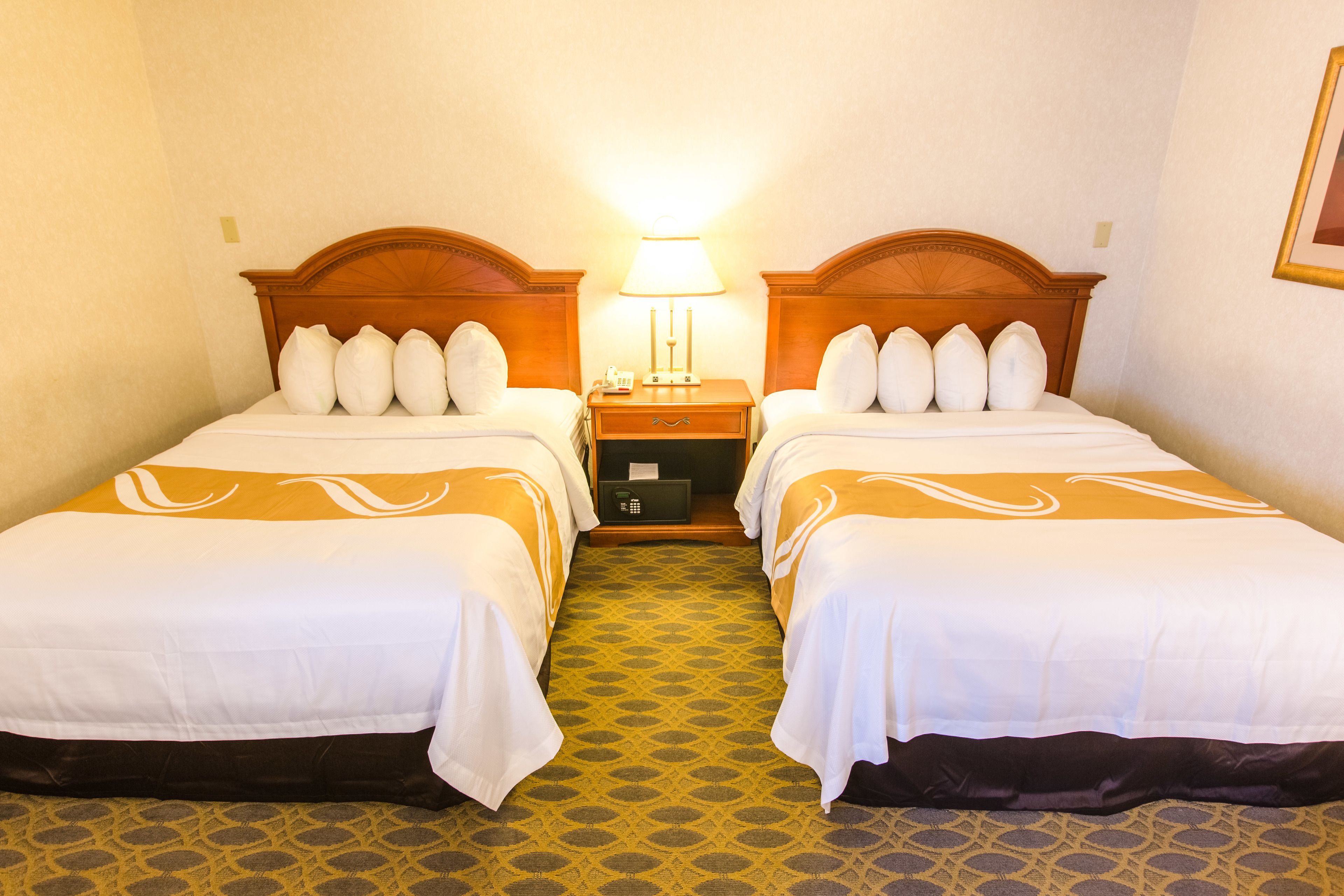 Une chambre d’hôtel avec deux lits, chacun étant équipé de draps blancs et d’un tapis à motifs. Une table de chevet avec une lampe se trouve entre les deux lits.