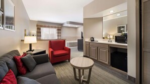 A compact hotel room with a sofa, a small round table, a red armchair, a kitchenette, and a large mirror. - Country Inn & Suites by Radisson, Freeport, IL (Freeport)