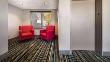 Un vestíbulo de hotel con alfombra a rayas, dos sillones rojos, una ventana con persianas y una puerta con el número.