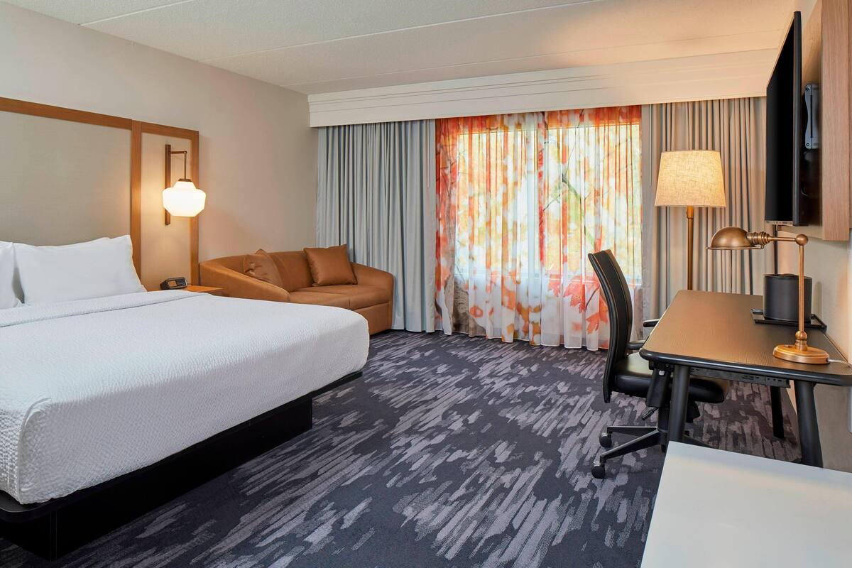 A hotel room with a large bed, a desk with a lamp, a chair, and a television on the wall.
