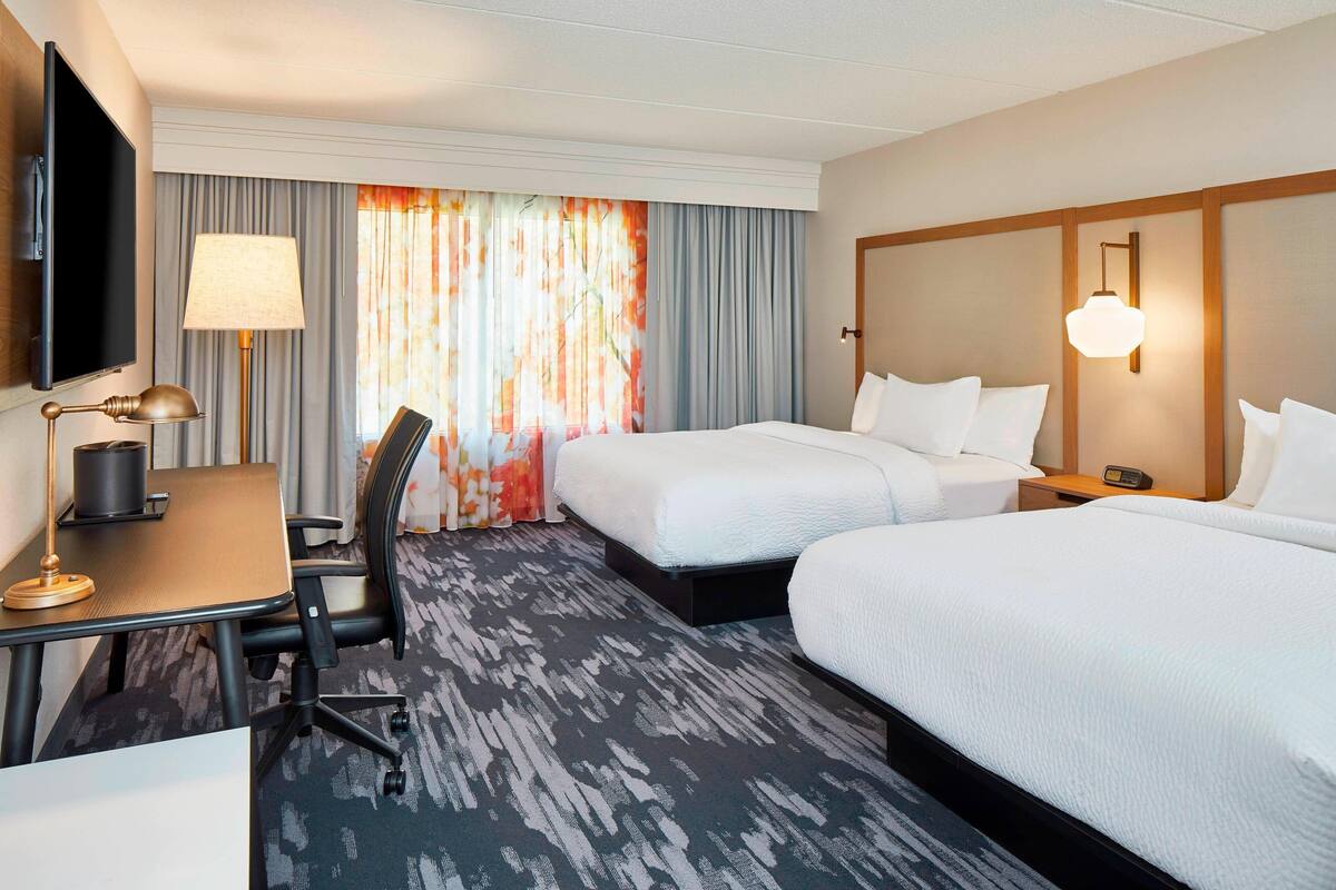 A hotel room with two beds, a desk with a lamp, a TV, and a large window with curtains.