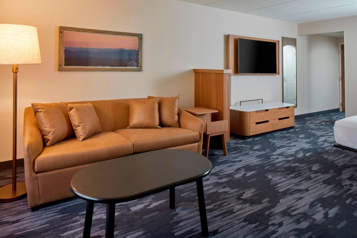 A hotel room with a sofa, a small table, a desk with drawers, a flat-screen TV, and a carpeted floor.
