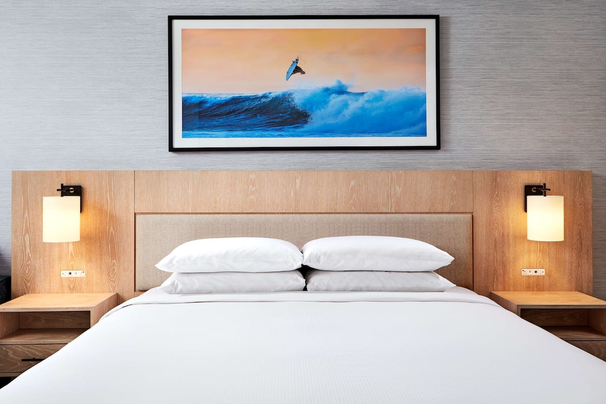Una habitación de hotel con una cama, dos mesitas de noche, un cabecero y un cuadro con una imagen de un surfista sobre una ola.