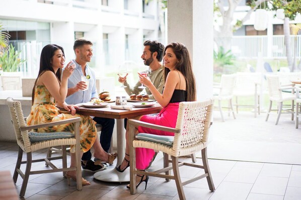 A group of people seated around a table in a bright, open-air dining area with wicker chairs and a tiled floor.