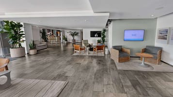 Un vestíbulo amplio con un diseño moderno, que cuenta con un gran espacio de estar con sofás y sillones, un televisor de pantalla plana en la pared y una variedad de plantas en macetas.
