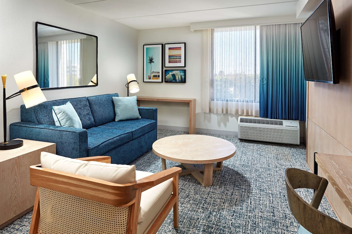 Una habitación de hotel moderna con un sofá azul, una mesa redonda de madera, una silla de mimbre, un televisor de pantalla plana y obras de arte en la pared.