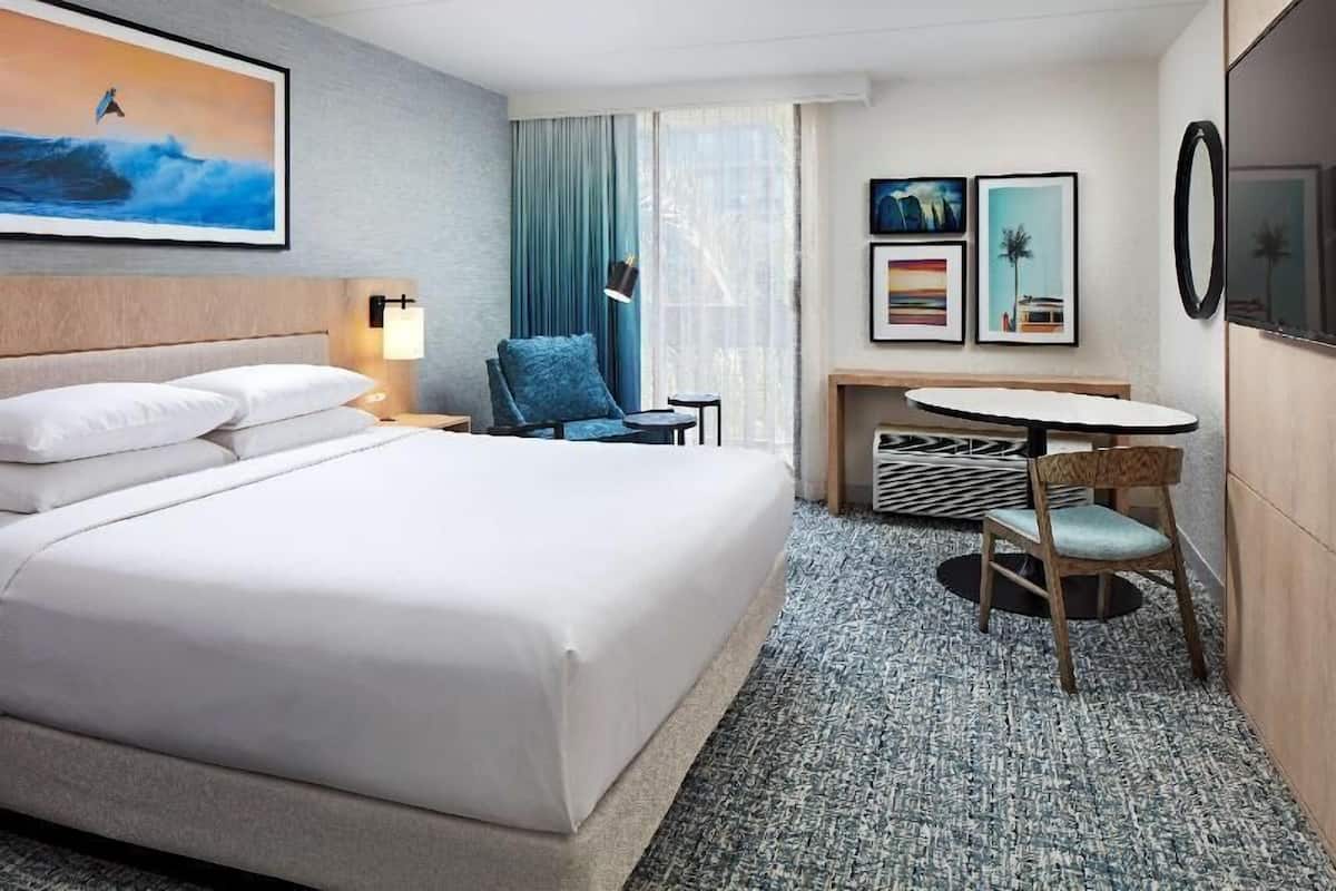 Habitación de hotel con una cama grande, un escritorio con silla, una mesita pequeña y un amplio ventanal con cortinas.
