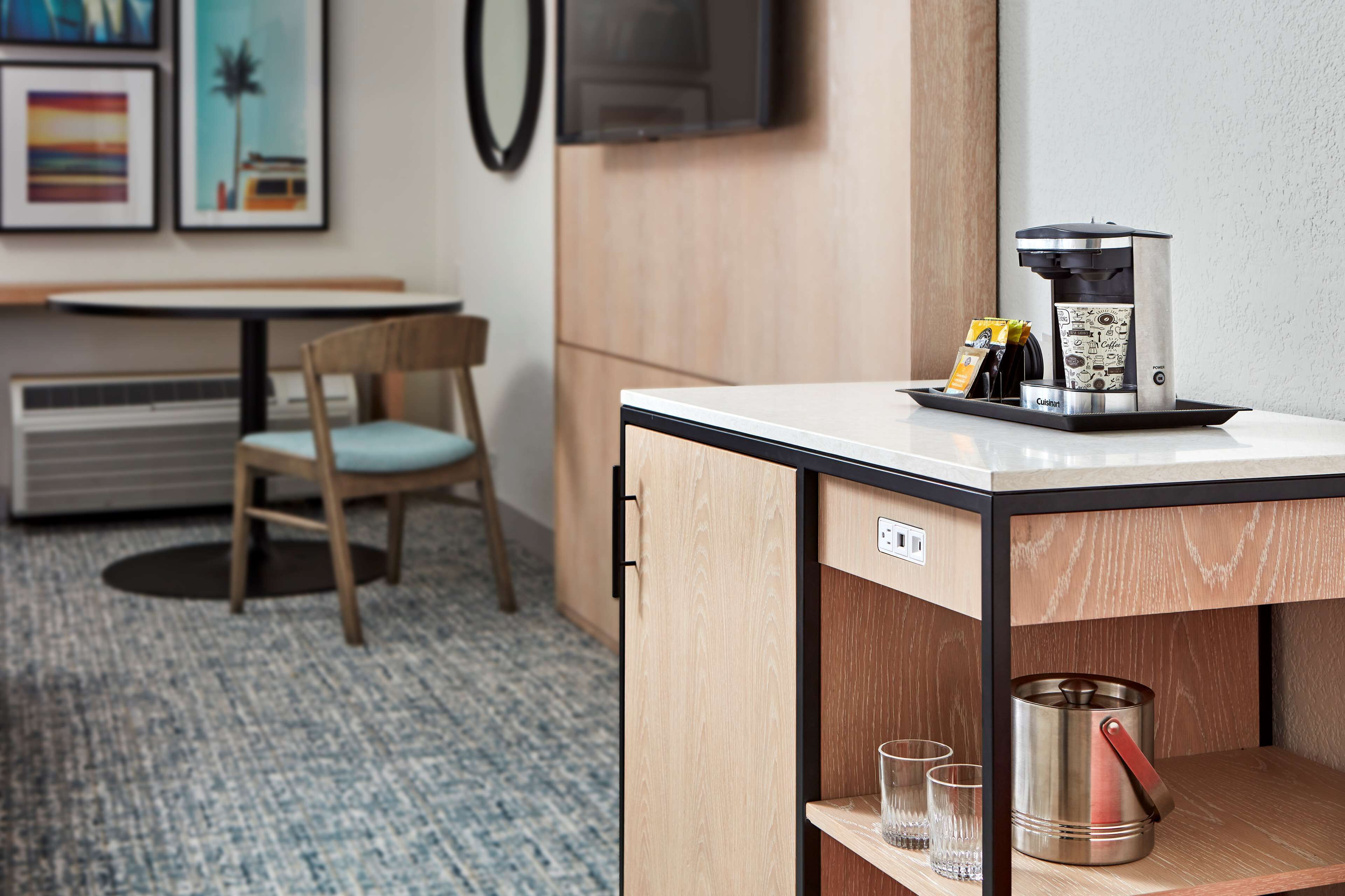 Une petite cuisine équipée comprenant une machine à café, une petite étagère avec des verres et un placard contenant une casserole.