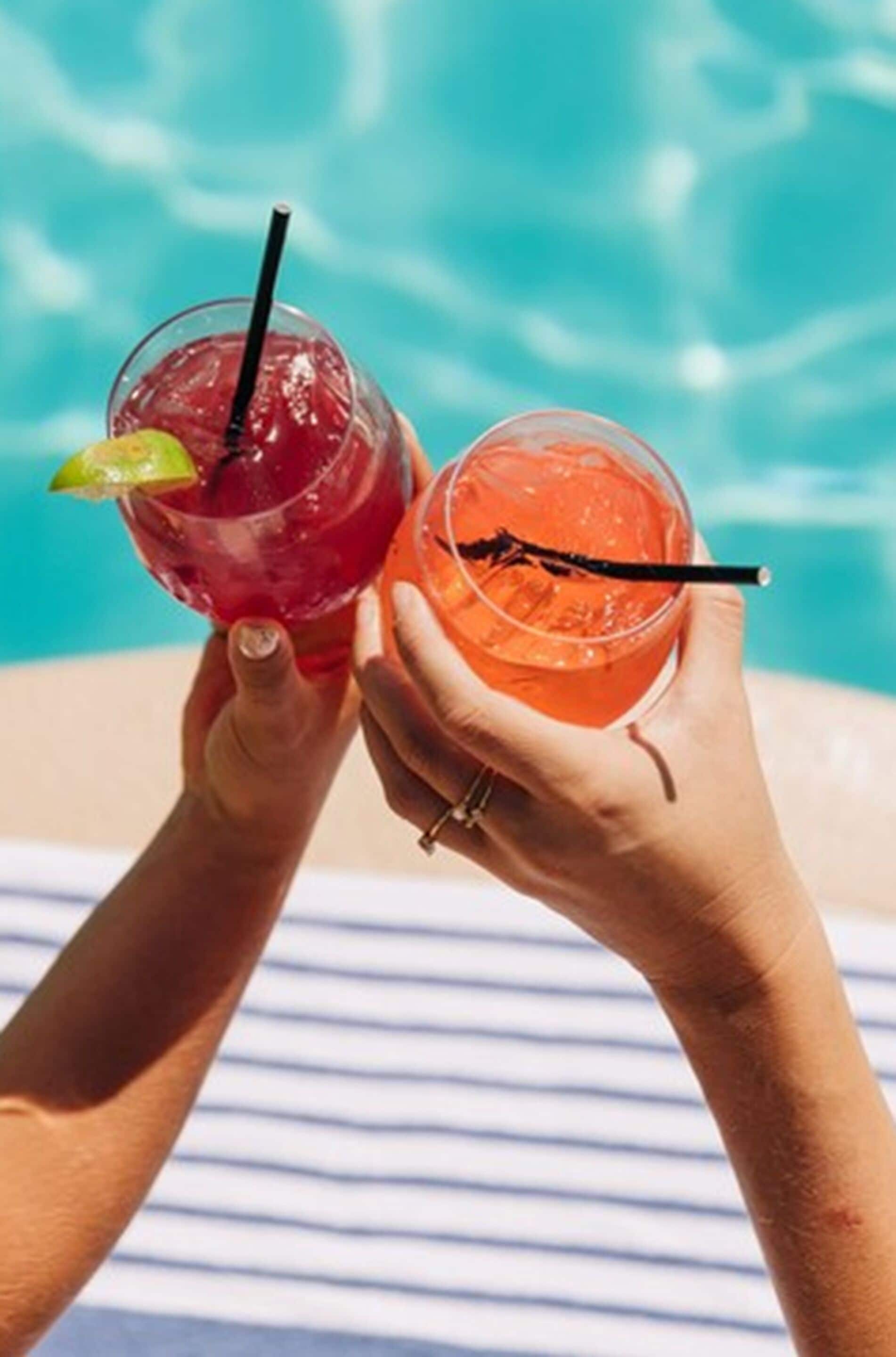 Deux mains tenant des verres à cocktail avec des pailles, l’un d’eux contenant un quartier de citron vert, près d’une piscine.