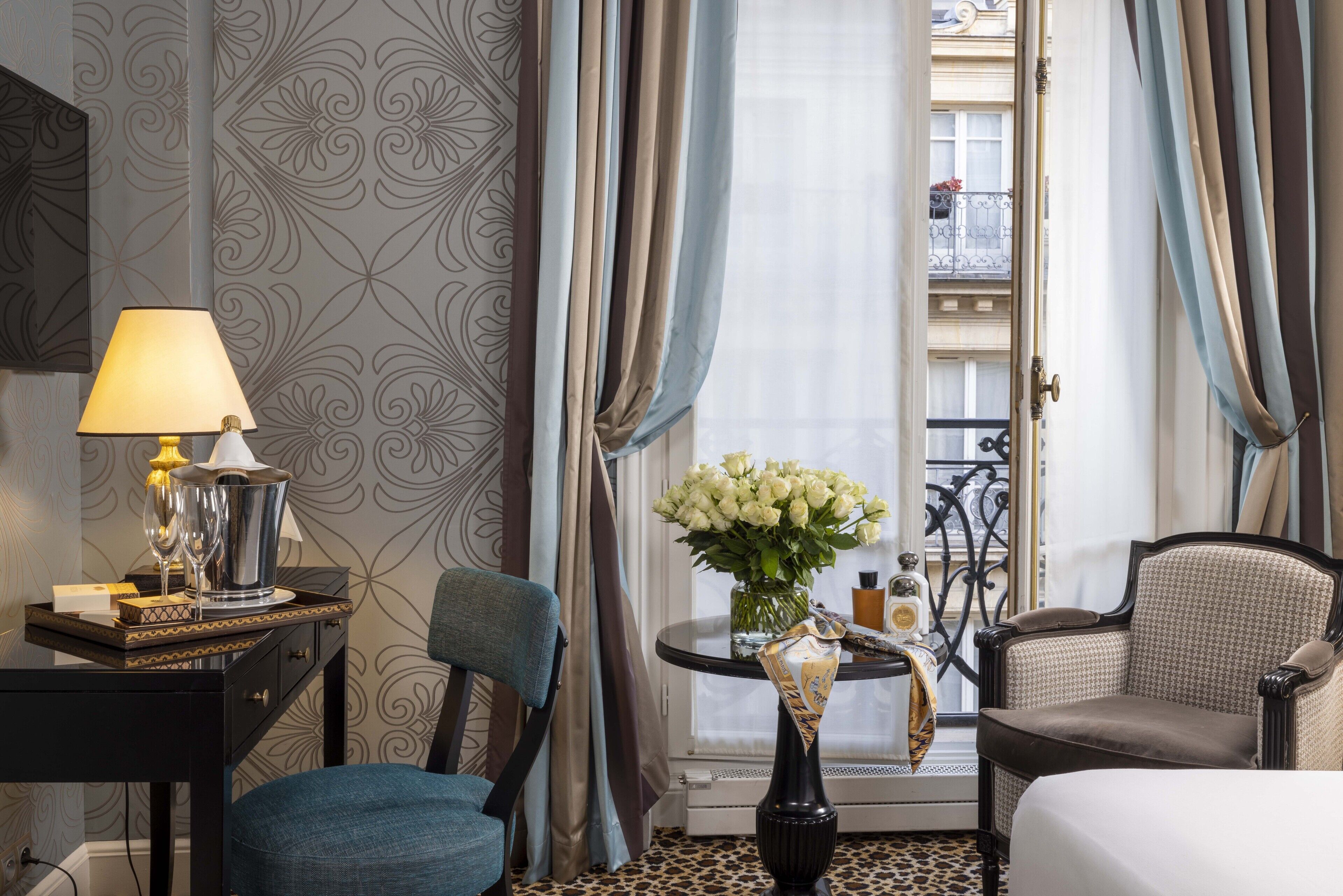 Une chambre d’hôtel avec un lit, un bureau doté d’une lampe, une chaise, une petite table ornée d’un vase de fleurs et une vue sur un balcon, visible à travers une fenêtre avec des rideaux.
