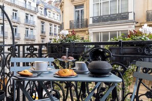 A table with a cup, a muffin, a teapot, and a tray with pastries on a balcony with wrought iron railings and buildings in the background. - West End Hotel (Paris)