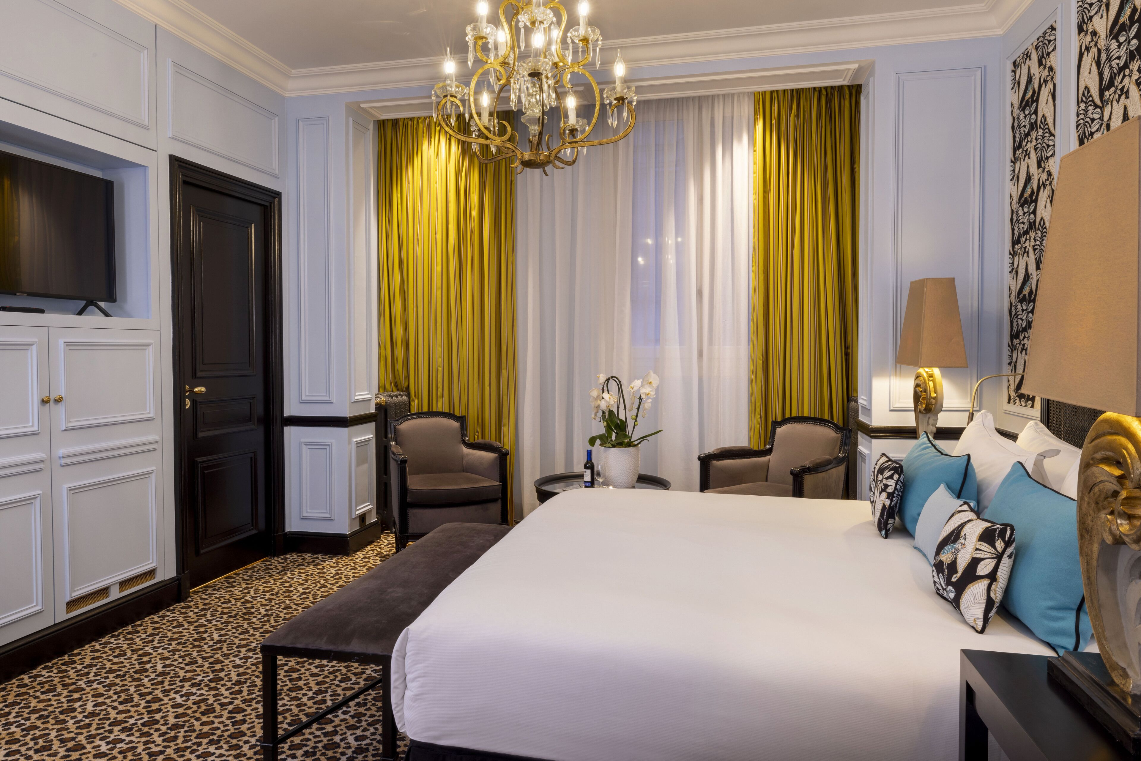 Una habitación de hotel con una cama grande, dos sillones, una mesita con un jarrón de flores, un televisor y una araña.
