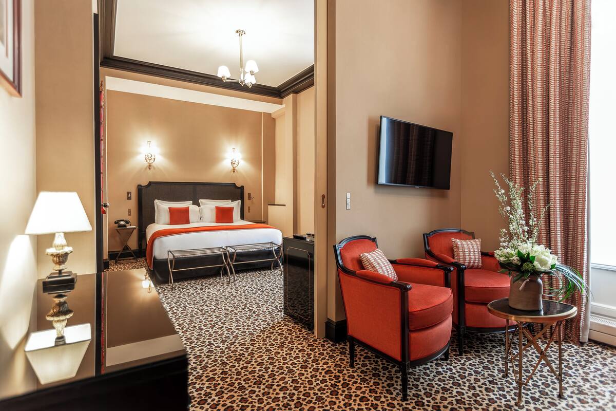 A hotel room with a bed, two red armchairs, a small table with a vase, a flat-screen TV, and a leopard print carpet.