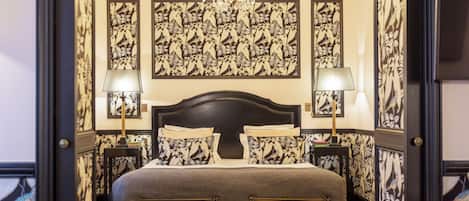 A bedroom with a large bed, two bedside tables with lamps, a chandelier, and floral wallpaper.