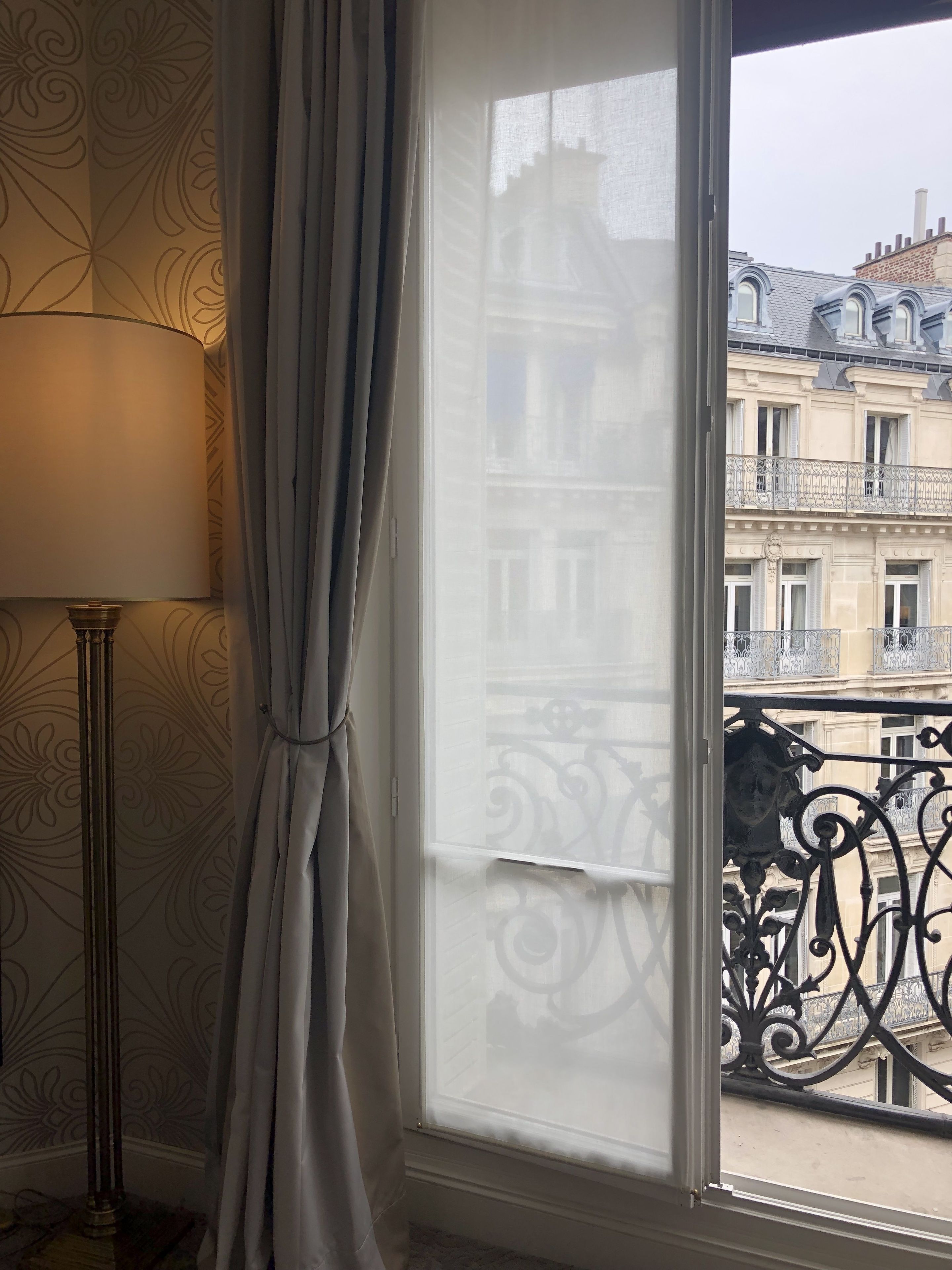 A room with a large window, a floor lamp, and a view of a building with balconies.