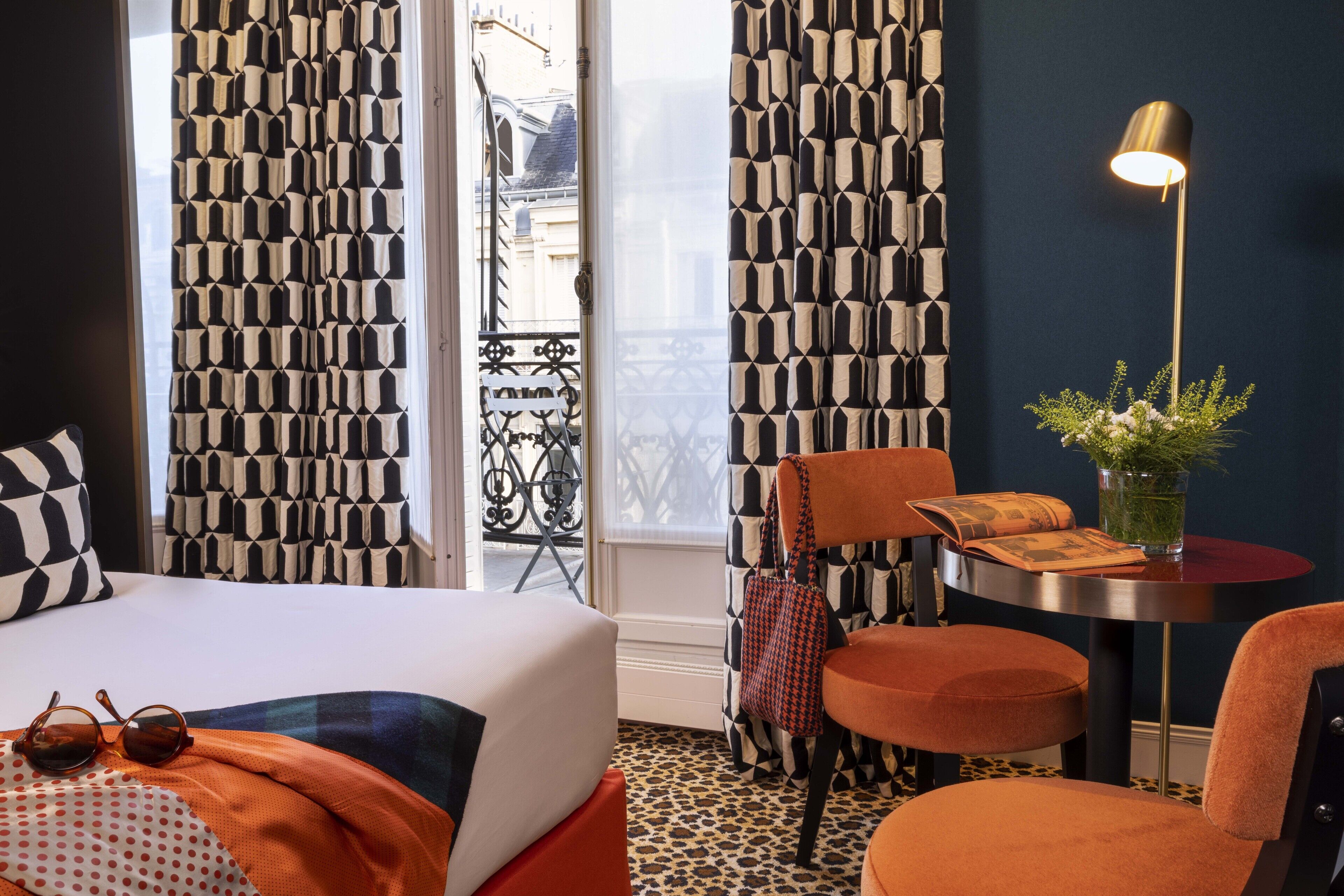 Una habitación de hotel con cama, una silla, una mesa con lámpara y cortinas con diseño geométrico.