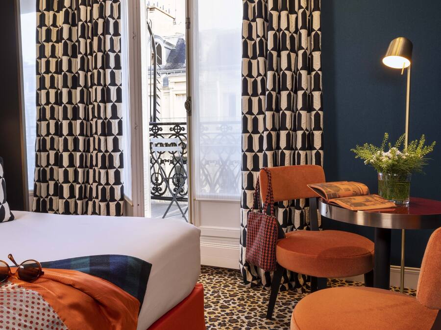 Una habitación de hotel con cama, una silla, una mesa con lámpara y cortinas con diseño geométrico.