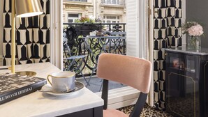 Una habitación con una mesa, una silla, una lámpara, una taza, un libro y vistas al balcón exterior.
