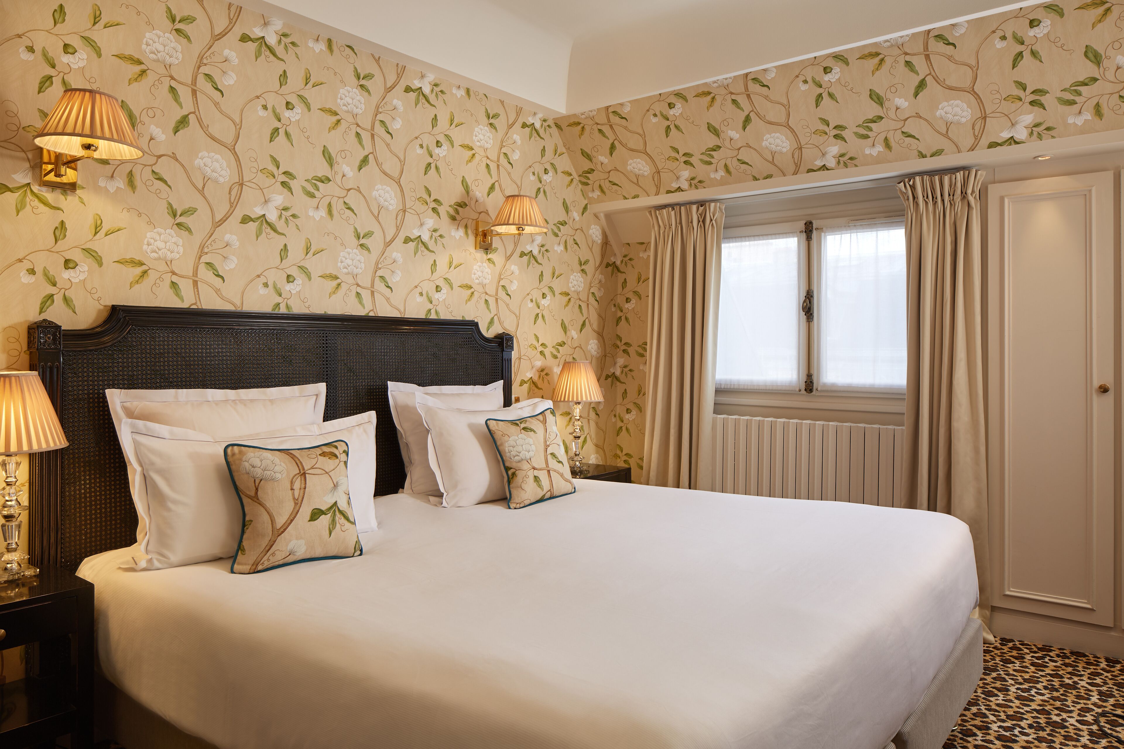 Una habitación de hotel con una cama grande, papel tapiz floral, dos lámparas de noche y una ventana con cortinas.