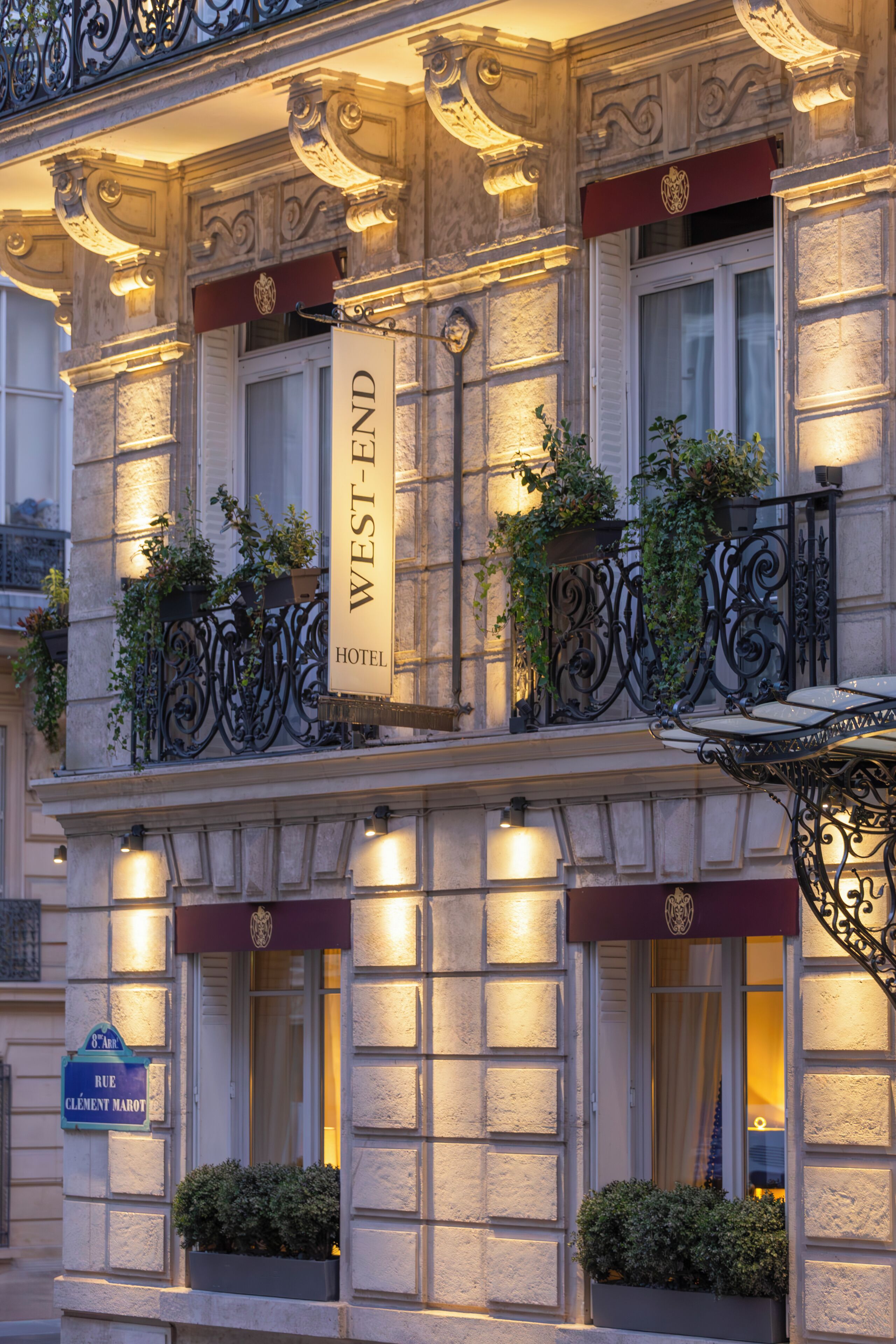 Fachada de hotel con un letrero que dice 'WEST-END HOTEL', barandillas de hierro decorativas y plantas en macetas.