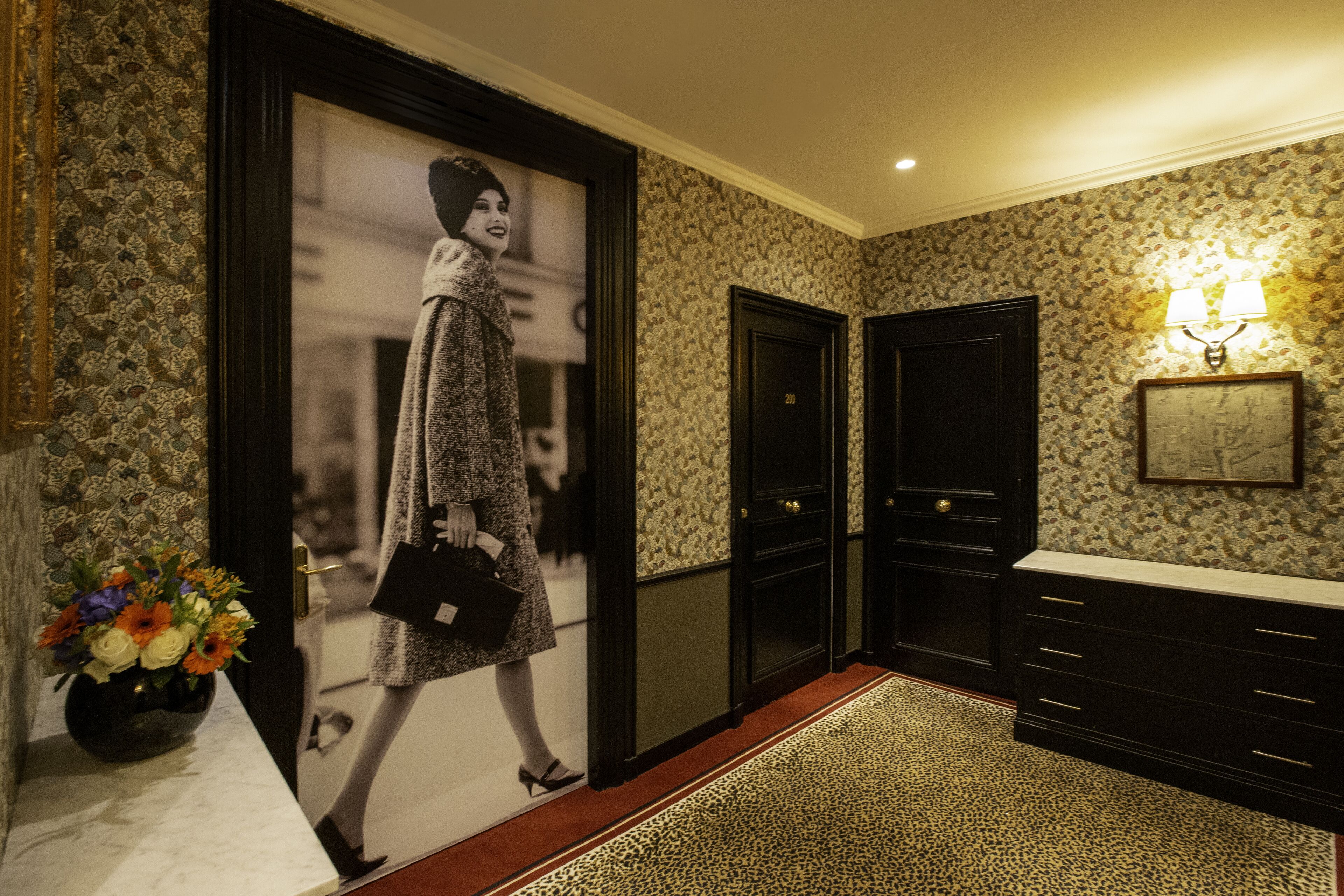 A hallway with patterned wallpaper, a black door, a wall-mounted light fixture, and a vase with flowers on a table.