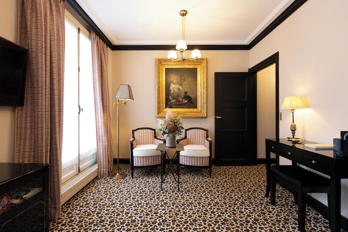 A hotel room with a checkered floor, a painting above a table, and a desk with a lamp.