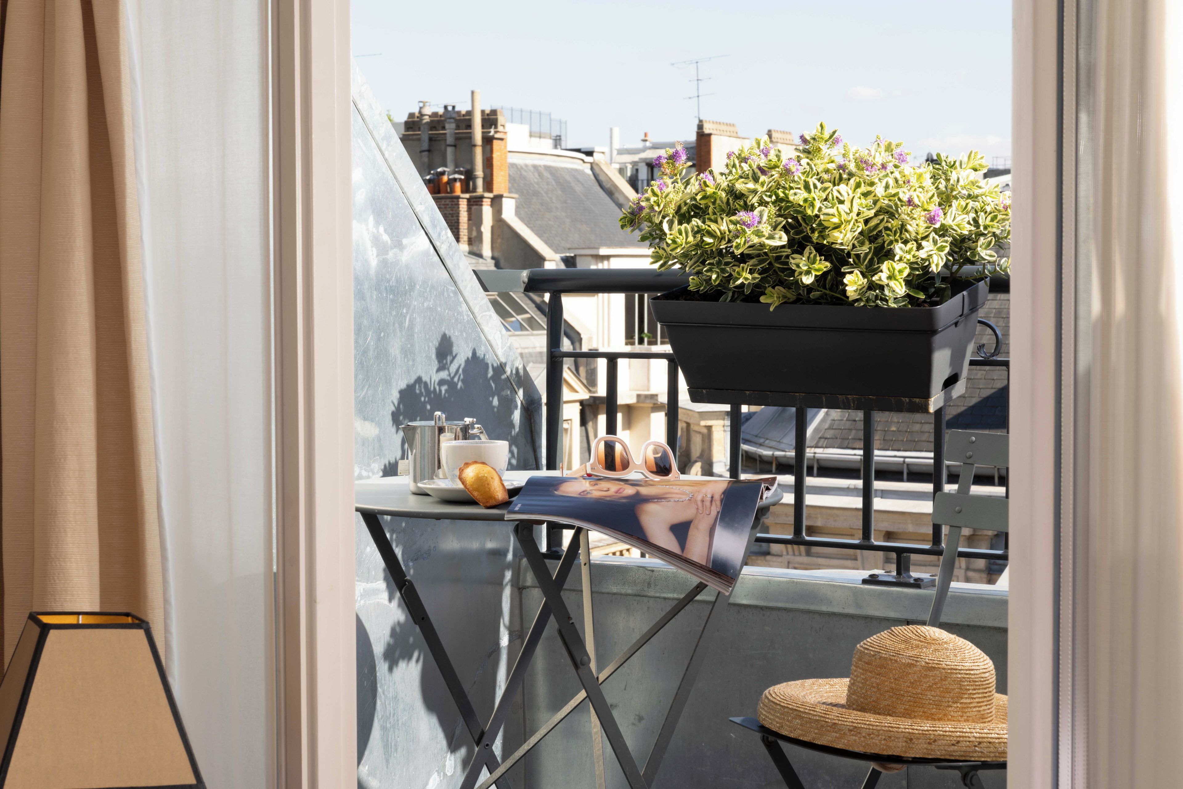 A balcony with a potted plant, a table with a cup, a pastry, and a magazine.