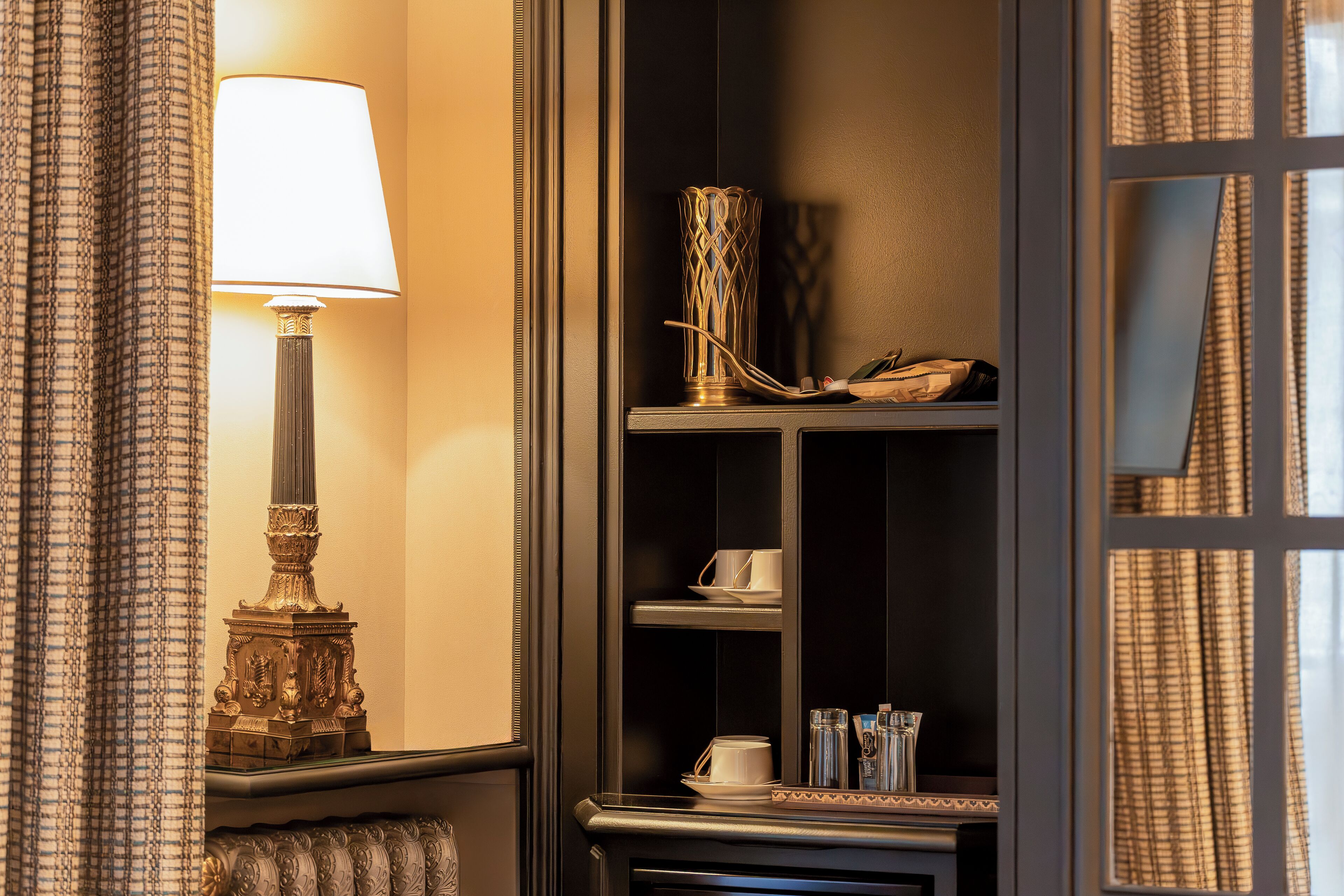 A corner of a hotel room with a lamp, a decorative vase, and a shelf with cups and a phone.